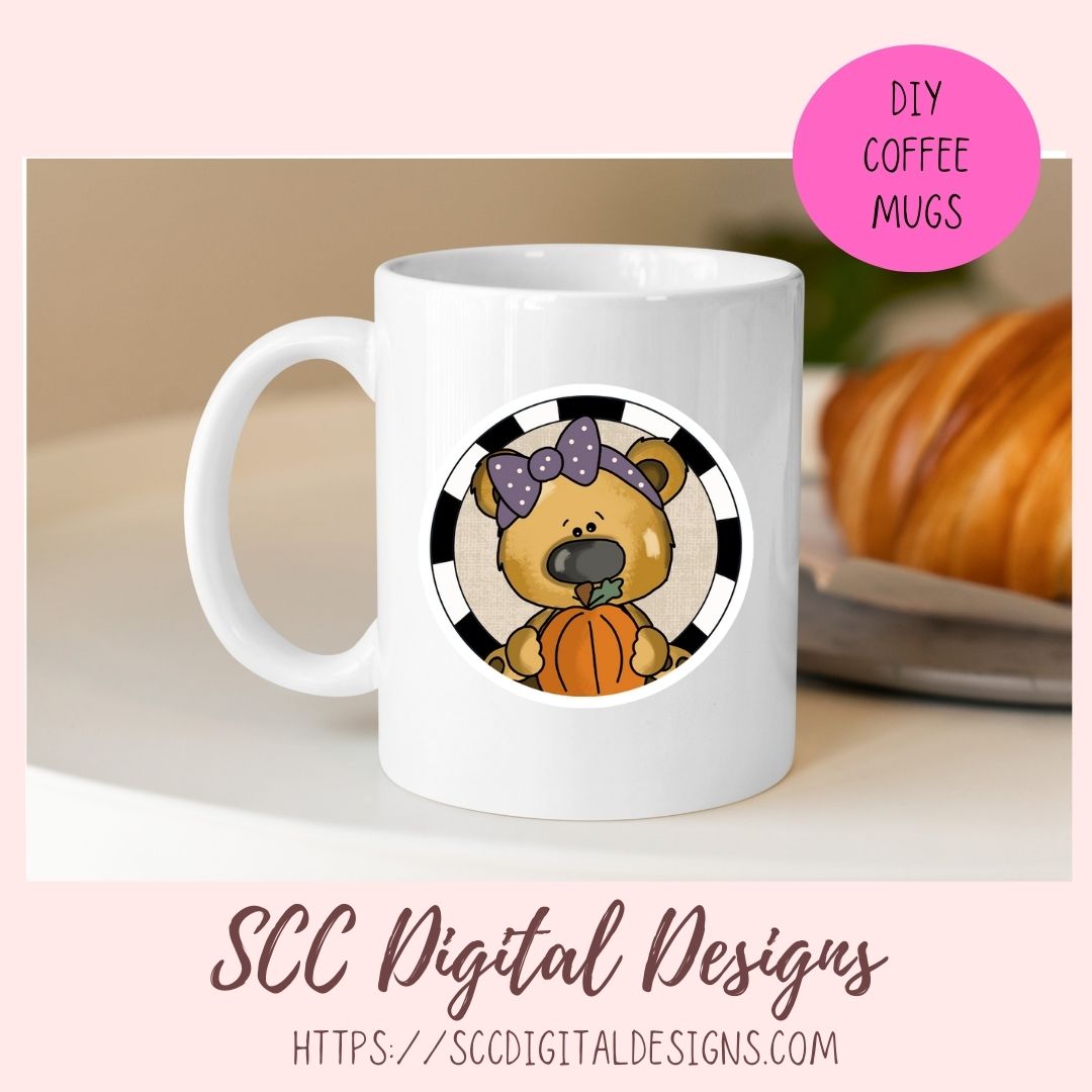 A white ceramic coffee mug with a circular cartoon design of a brown bear holding an orange pumpkin, set against a black and white striped background. The mug is displayed on a light surface with a blurred croissant in the background.