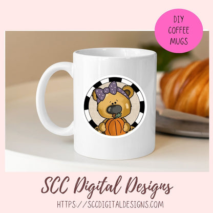 A white ceramic coffee mug with a circular cartoon design of a brown bear holding an orange pumpkin, set against a black and white striped background. The mug is displayed on a light surface with a blurred croissant in the background.