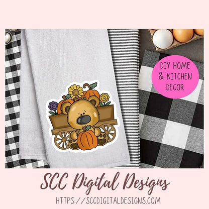 A styled flat-lay mockup showing a die-cut sticker of a cartoon bear in a wagon with pumpkins and flowers, placed on a light gray towel alongside black-and-white buffalo plaid and striped fabrics. A pink circle reads 'DIY HOME & KITCHEN DECOR,' and the bottom text says 'SCC Digital Designs' with a website URL.