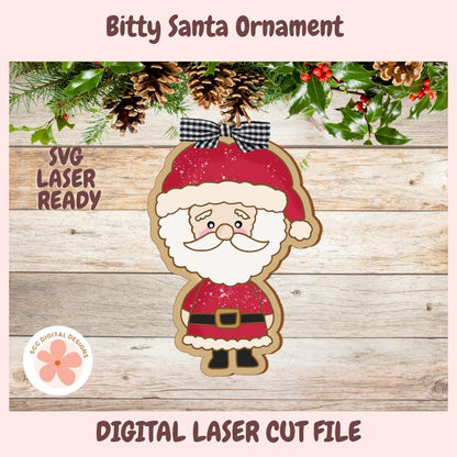 Digital preview of a Christmas ornament design featuring a cute Santa Claus figure with a red hat, white beard, black-and-white bow, and red outfit on a rustic wood background with pinecones and holly. Text reads 'Bitty Santa Ornament,' 'SVG LASER READY,' and 'DIGITAL LASER CUT FILE.