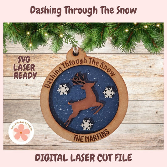 Digital preview of a Christmas ornament design with a brown reindeer silhouette on a dark blue circular background, surrounded by white snowflakes and a natural wood border. The text reads 'Dashing Through The Snow' and 'THE MARTINS.