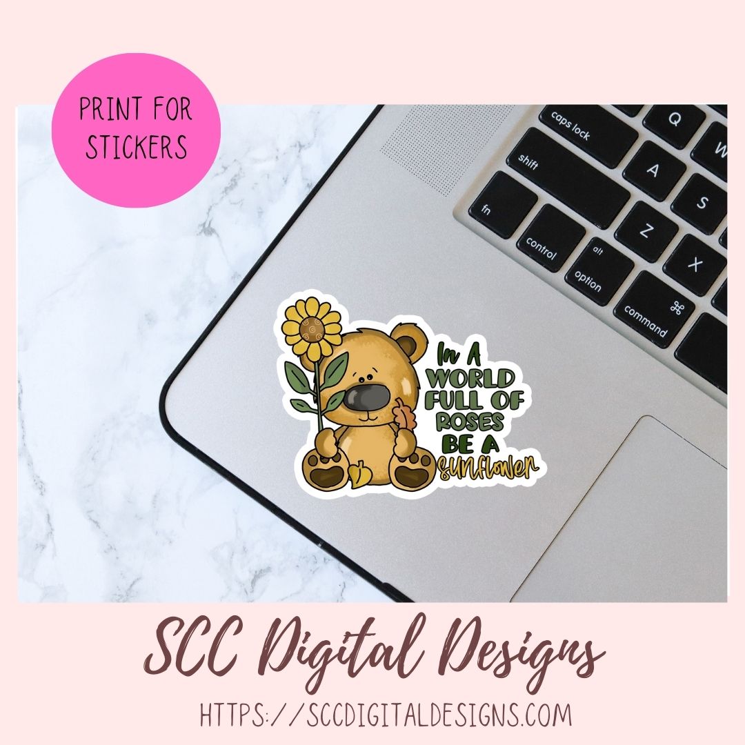 A sticker design shown on a laptop trackpad over a marble surface, with a pink circle reading 'PRINT FOR STICKERS' and text at the bottom for 'SCC Digital Designs'. The sticker features a cartoon teddy bear holding a sunflower with the text 'In A WORLD FULL OF ROSES BE A RUMOR'.