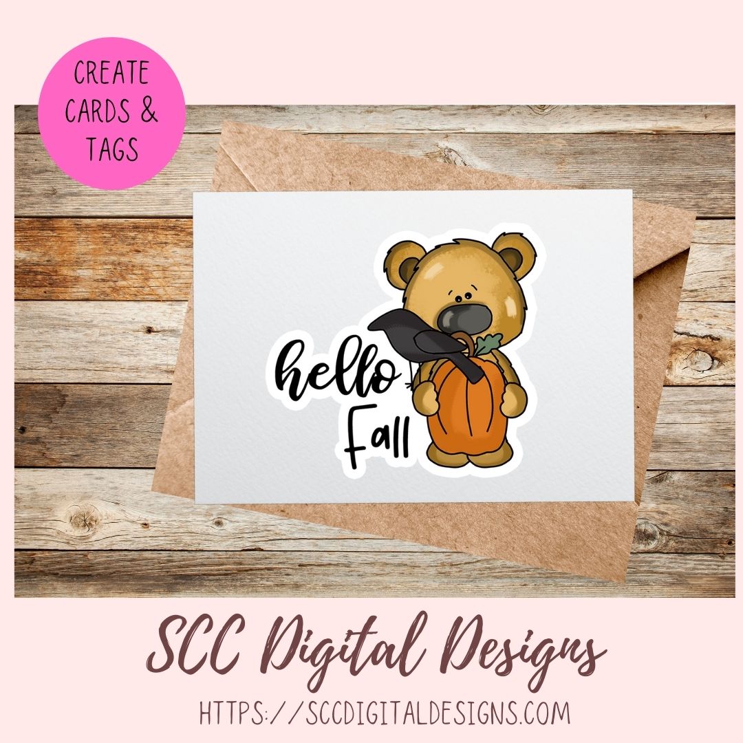 A digital design preview showing a white card with a cartoon bear holding an orange pumpkin and a black hat, next to a brown kraft envelope on a wood background. A pink circle says 'CREATE CARDS & TAGS' and the bottom text reads 'SCC Digital Designs' with a website URL.