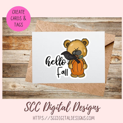 A digital design preview showing a white card with a cartoon bear holding an orange pumpkin and a black hat, next to a brown kraft envelope on a wood background. A pink circle says 'CREATE CARDS & TAGS' and the bottom text reads 'SCC Digital Designs' with a website URL.