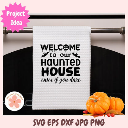 A white waffle-knit kitchen towel hanging on a metal rack, printed with the phrase 'WELCOME to our HAUNTED HOUSE enter if you dare' in black text with small bat and skull motifs. The image includes decorative pumpkins at the bottom right, a pink 'Project Idea' badge at top left, and file-format text at the bottom.