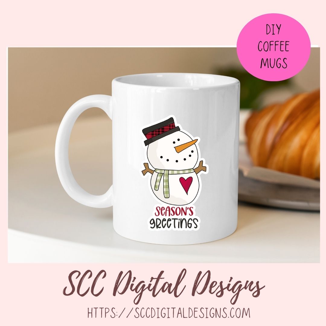 A white ceramic coffee mug with a winter holiday design featuring a cartoon snowman wearing a plaid top hat and scarf, with the text 'SEASON'S GREETINGS' and a red heart. The mug is presented in a mockup setting with a pink background, a blurred croissant in the background, and a pink circle badge that reads 'DIY COFFEE MUGS'.