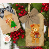 Two Christmas-themed teddy bear ornaments on brown paper bags with ribbons, surrounded by greenery and red berries.