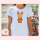 White t-shirt with a cartoon reindeer design on a wooden background with red berries.