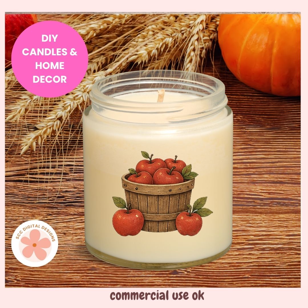 A lit soy candle in a clear glass jar with a cream-colored wax and a printed label of red apples in a wooden basket. The jar is set on a rustic wooden surface with decorative wheat stalks and a pumpkin in the background.