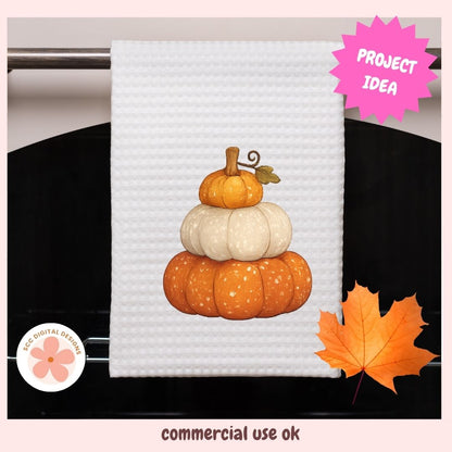 A white waffle-weave kitchen towel hanging on a metal rack, featuring a stacked autumn pumpkin design in orange and cream. The image includes a pink 'PROJECT IDEA' starburst, a circular 'ECO-DIGITAL DESIGNS' logo, and a decorative orange maple leaf on a light pink background with 'commercial use ok' text at the bottom.