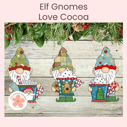 Three festive elf gnome figures decorated with colorful winter hats, red-and-white striped hats, white beards, and green pants, each holding a hot cocoa mug filled with marshmallows or a Christmas tree ornament. The scene is set on a rustic whitewashed wood surface with evergreen branches and red berries across the top, under the text 'Elf Gnomes Love Cocoa.