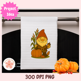 White towel with a cartoon gnome holding pumpkins and corn, displayed on a towel rack with pumpkins below.