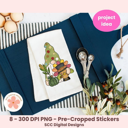 A styled flat-lay showing a white fabric napkin with a colorful Thanksgiving-themed gnome illustration, placed on navy blue paper with measuring cups, eggs, and olive branches around it. Pink text badges read 'project idea' and '8 - 300 DPI PNG - Pre-Cropped Stickers' with 'SCC Digital Designs' at the bottom, indicating this is a digital design preview rather than a straightforward physical product photo.