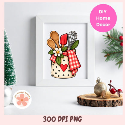 A framed kitchen utensil illustration displayed on a white surface with holiday props. The artwork shows a beige polka-dot jar filled with wooden spoons, a red spatula, a metal whisk, green leaves, a white flower, and a red gingham cloth with a red heart.