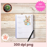 Notebook with a decorative sticker of a bunny holding a teddy bear on a wooden surface, with a pen and small plant.