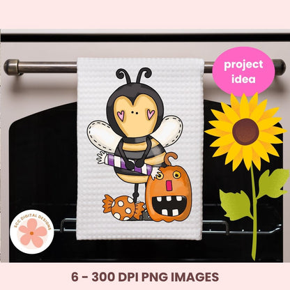 A white kitchen towel hanging on an oven rack, printed with a cute cartoon bee wearing a purple-and-white striped shirt and holding a pumpkin, with candy accents. The image includes a pink 'project idea' badge, a sunflower graphic, a circular '6 - 300 DPI PNG IMAGES' label, and a pink background border.