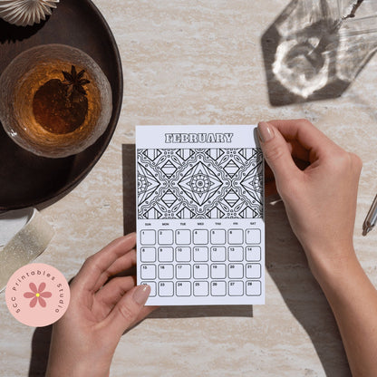 Person holding a February calendar with mandala design on a table with a drink and decorative items.