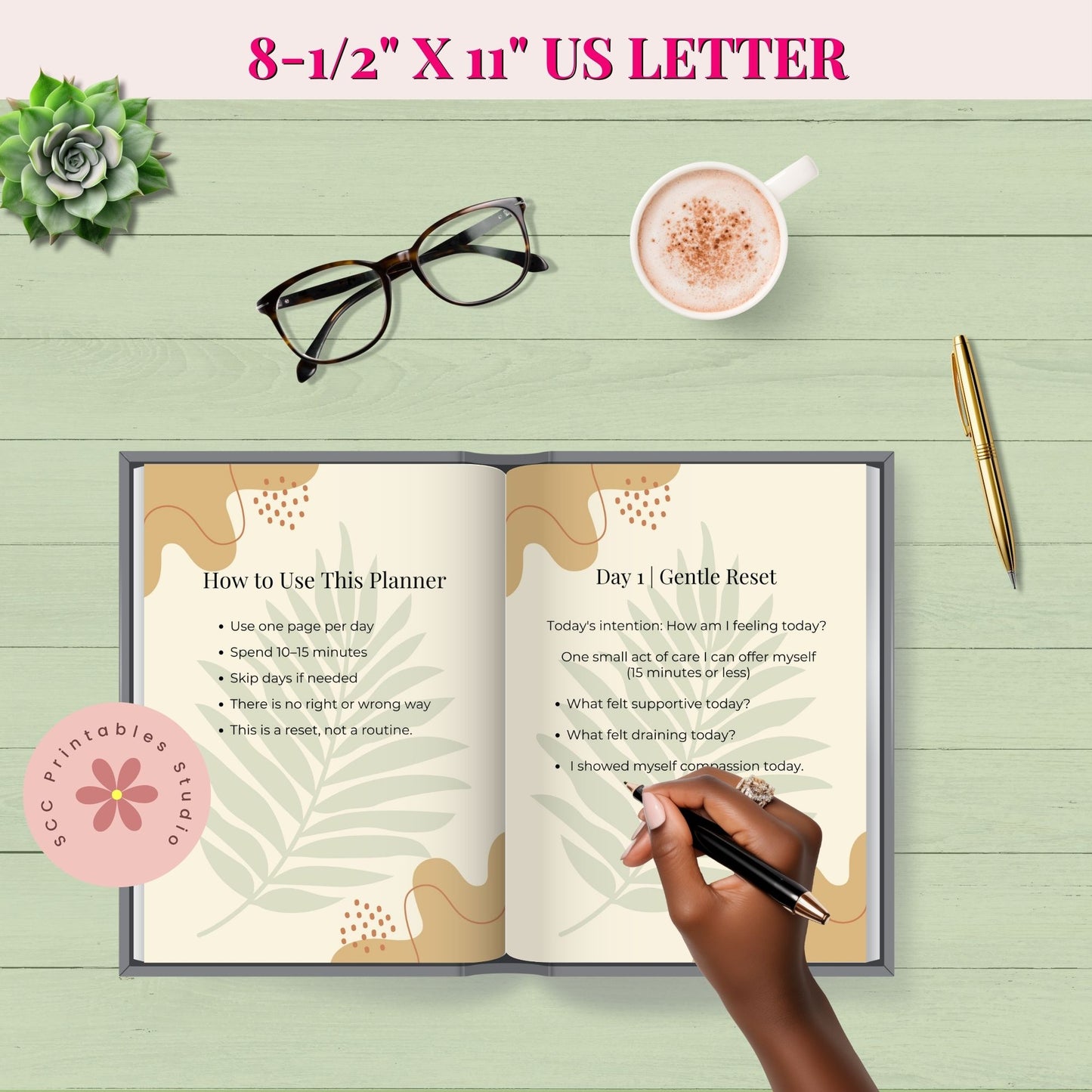 Open planner on a wooden surface with a hand holding a pen, a cup of coffee, glasses, and a succulent plant.