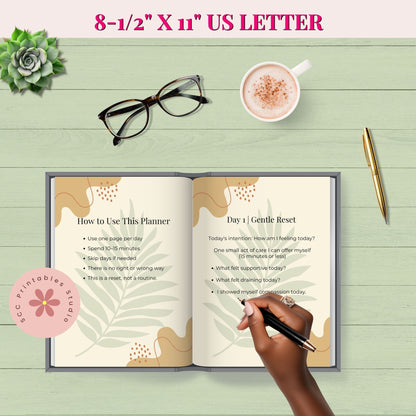 Open planner on a wooden surface with a hand holding a pen, a cup of coffee, glasses, and a succulent plant.