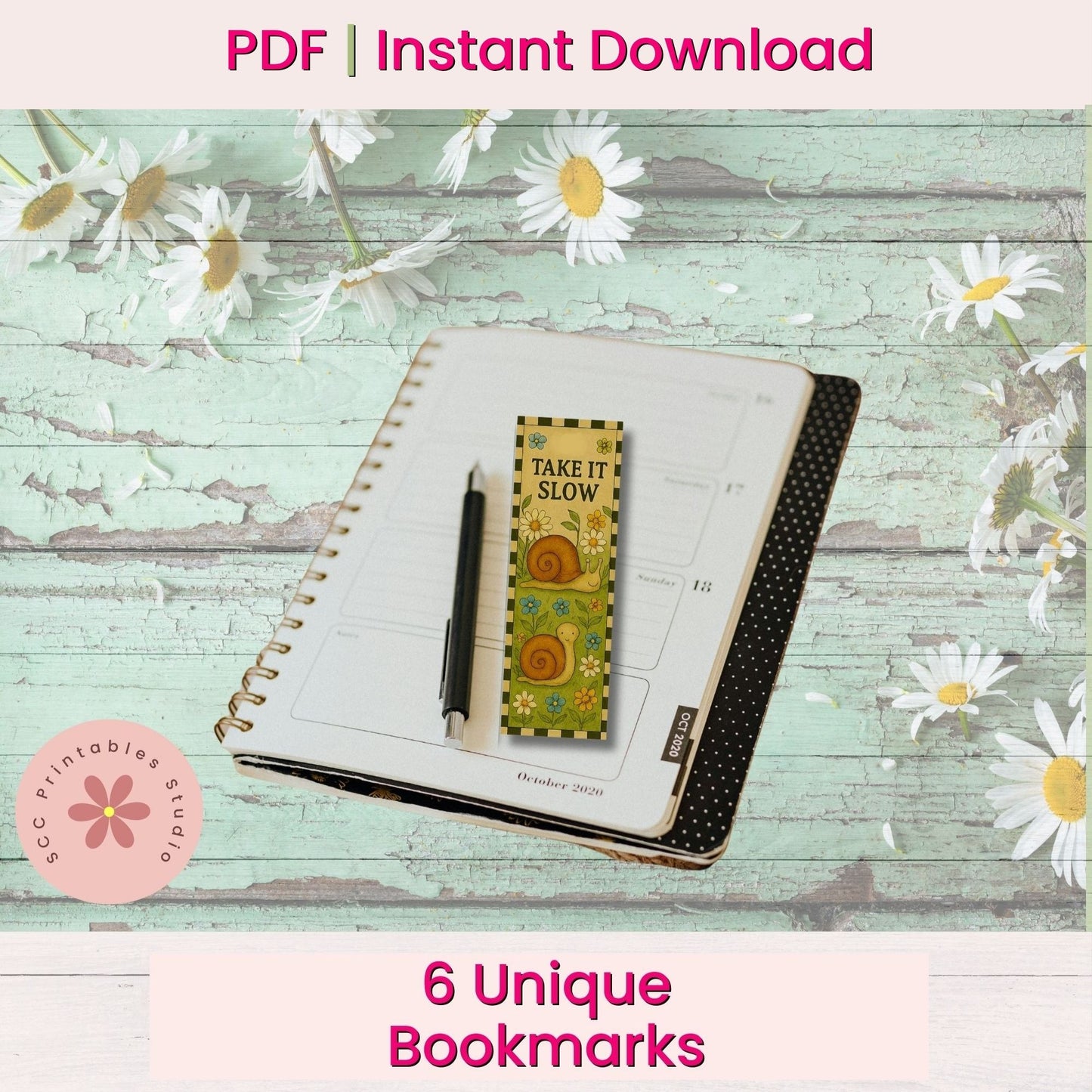 A styled flat-lay mockup showing a decorative bookmark with a yellow and green sun design reading 'TAKE IT SLOW' placed on an open notebook, with a black pen beside it. The scene is set on a distressed light green wood background with white daisies.