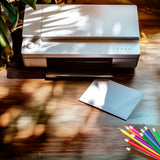 Laser printer on a wooden surface with a piece of paper and colored pencils.