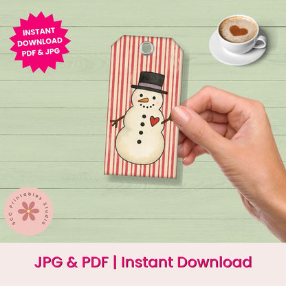 A hand holds a rectangular printed tag featuring a snowman illustration on a red-and-white striped background. The scene is styled like a desk setup with a cup of coffee in the upper right and text reading 'INSTANT DOWNLOAD PDF & JPG' and 'JPG & PDF | Instant Download Download.