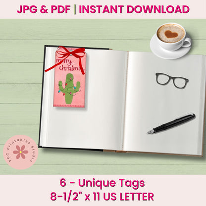 A promotional mockup for printable Christmas tags, showing an open notebook with a pink tag attached on the left page. The tag has a red ribbon bow and a green cactus illustration with the words 'merry christmas.