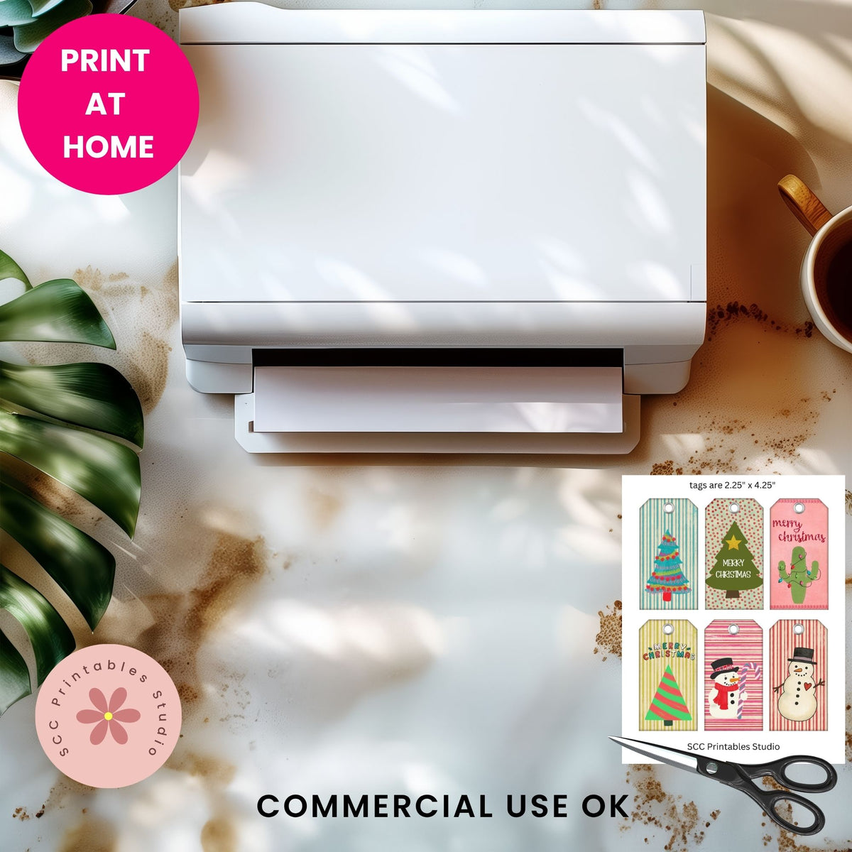 A white desktop printer shown on a marble surface with a pink 'PRINT AT HOME' badge, a pink 'SCC Printables Studio' logo, a sample holiday print sheet, and scissors nearby.