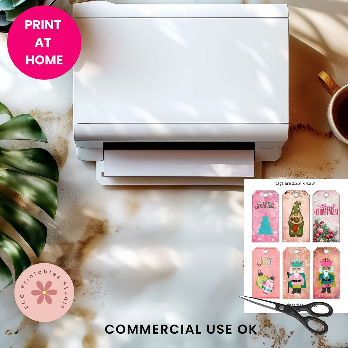 A white home printer shown from above on a marble-like surface with a coffee cup, green leaves, and a 'PRINT AT HOME' badge. A small inset shows colorful Christmas-themed printable tags and scissors, with text reading 'COMMERCIAL USE OK' and 'SCC Printables Studio'.