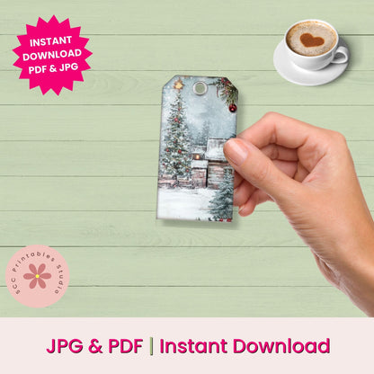 A hand holds a printed Christmas gift tag featuring a snowy winter village scene. The image includes promotional text reading 'Instant Download PDF & JPG' and 'JPG & PDF | Instant Download,' along with a cup of coffee with a heart-shaped latte on a light wood background.